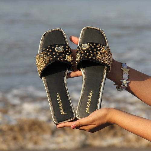 Black sandals with gold embellishments held by a person against a neutral background