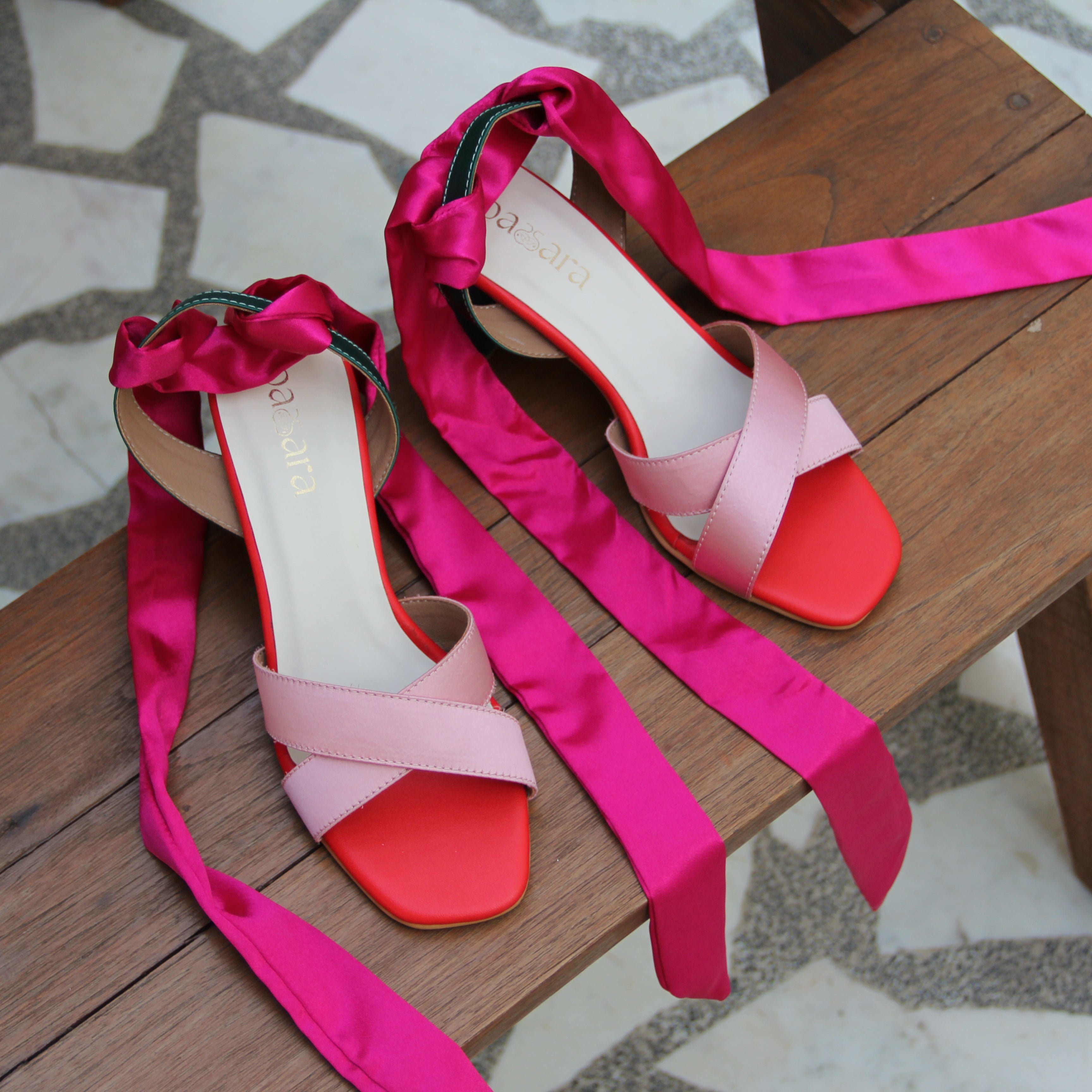 Pair of pink and red sandals with ribbon straps on a wooden bench.