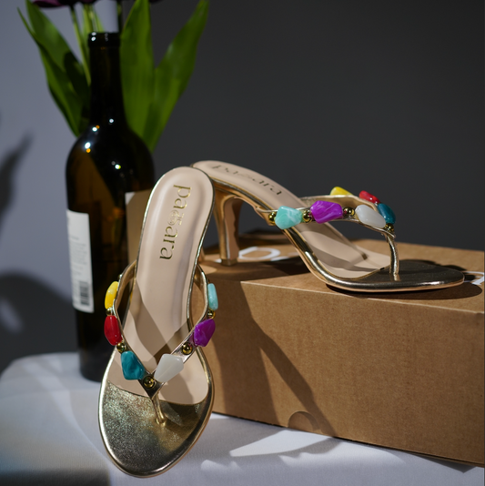 Gold sandals with colorful beads on a table with a box and flowers in the background