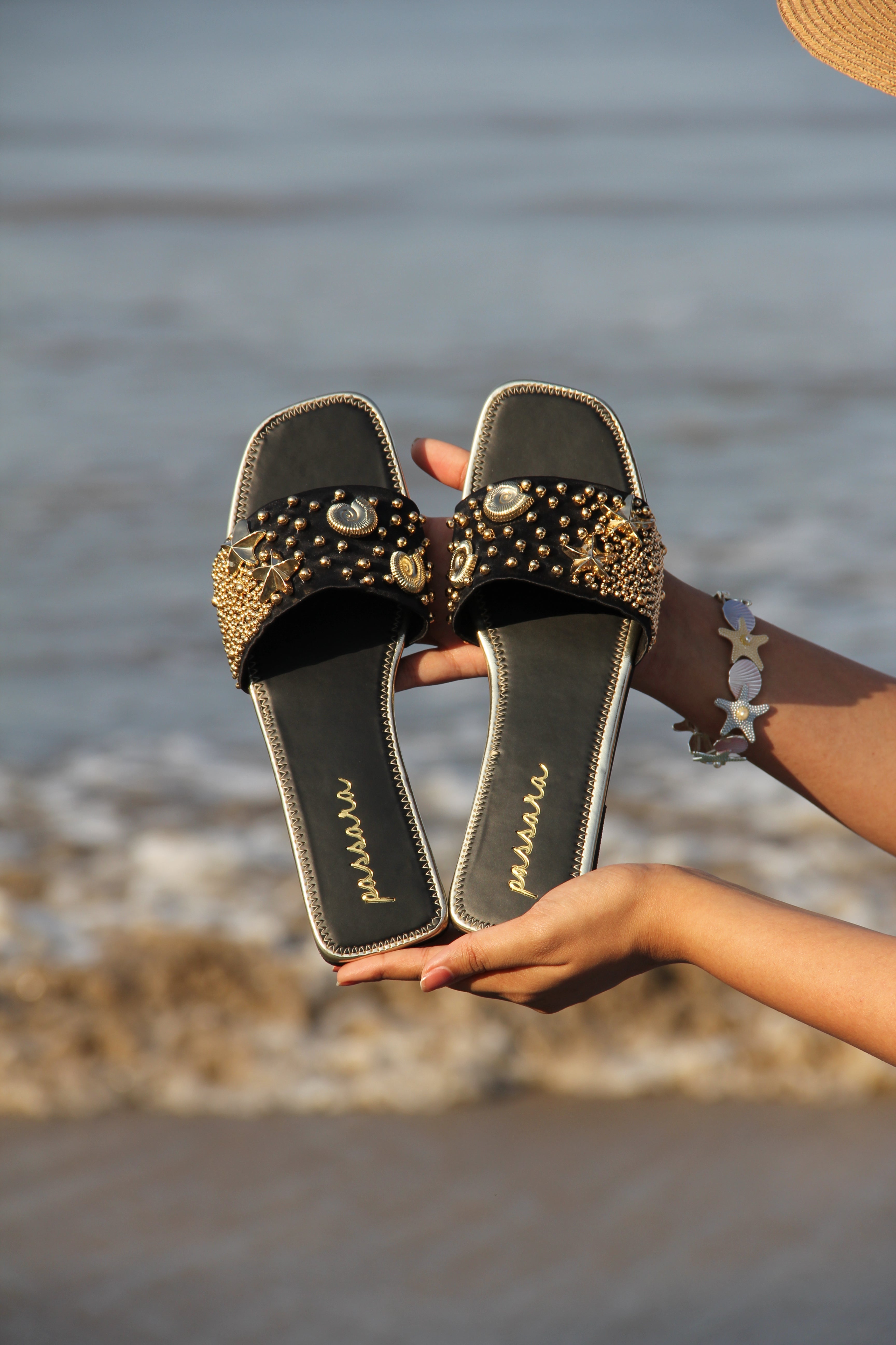 Black sandals with gold embellishments held by a person against a neutral background