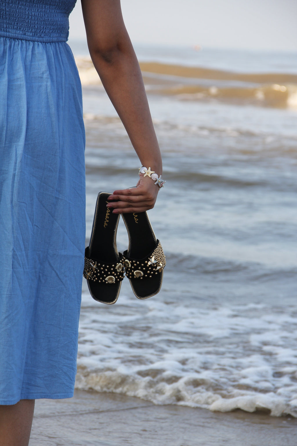 Person holding black high-heeled shoes with gold embellishments by a beach.