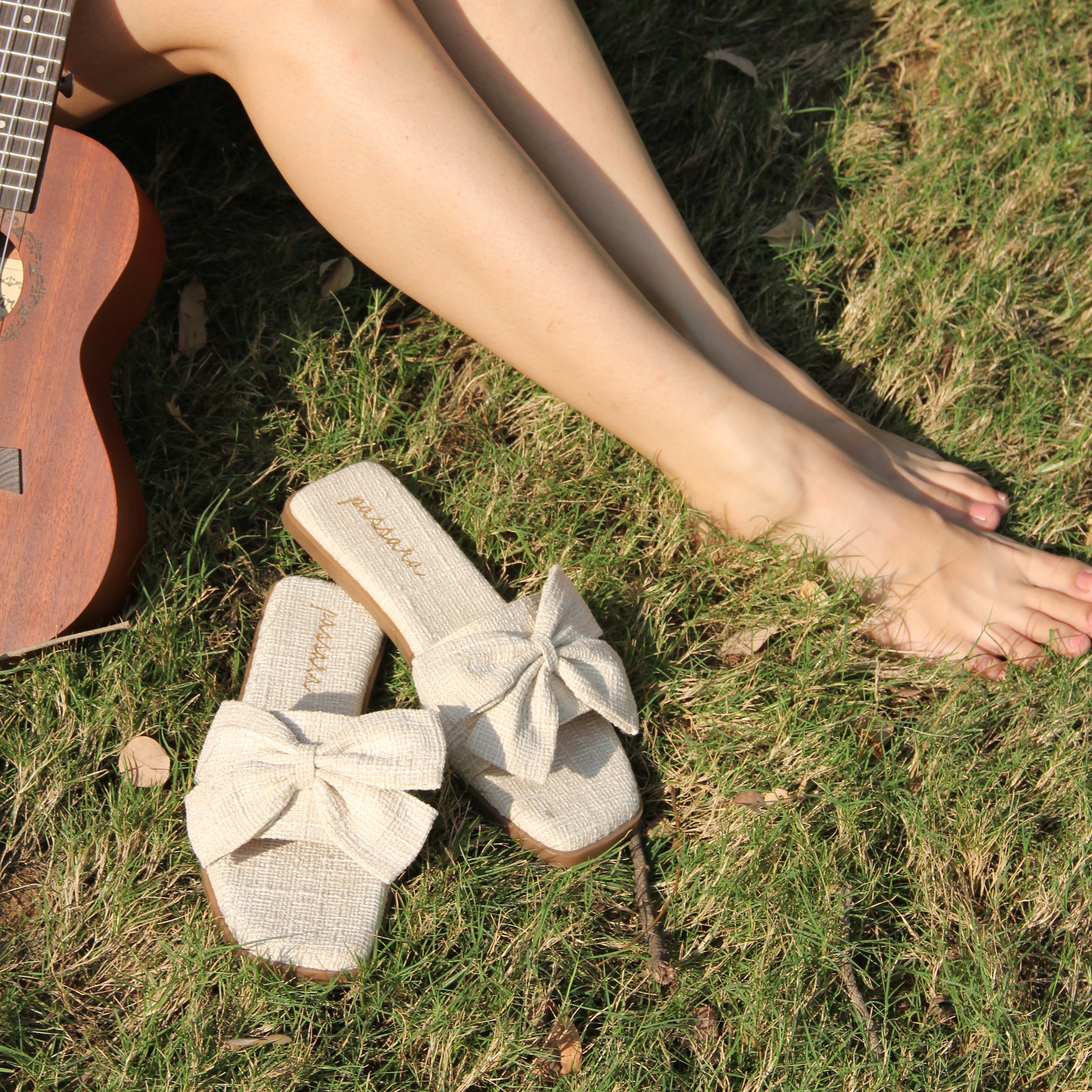 Person sitting on grass with legs crossed, wearing white sandals, holding a guitar, and holding red flowers.