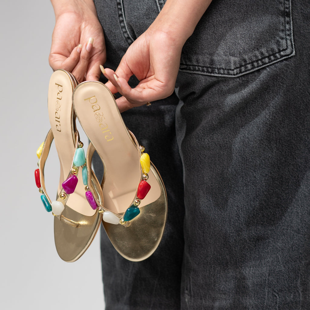 Gold sandals with colorful beads held by a person wearing dark jeans on a gray background