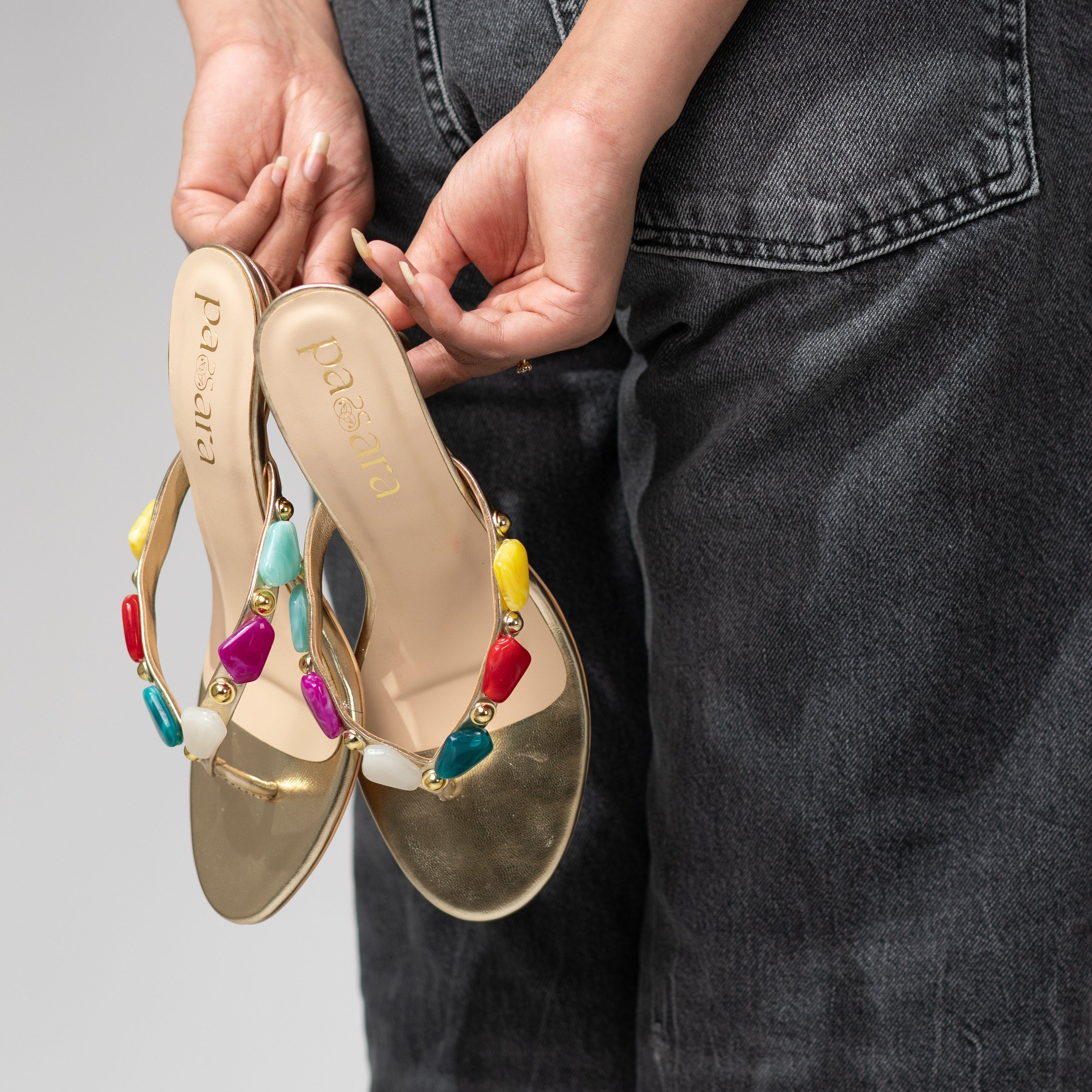 Gold sandals with colorful beads held by a person wearing dark jeans on a gray background